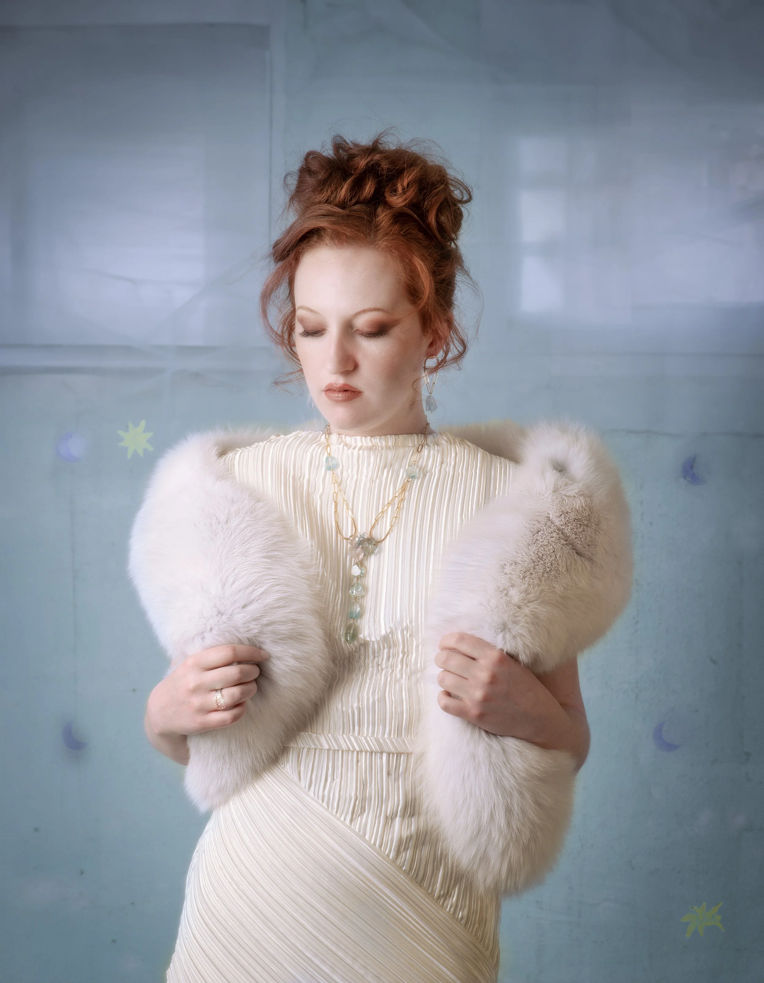 Portrait of a red-haired woman in a white pleated dress and fur stole, wearing delicate layered jewelry against a dreamy pale blue background.