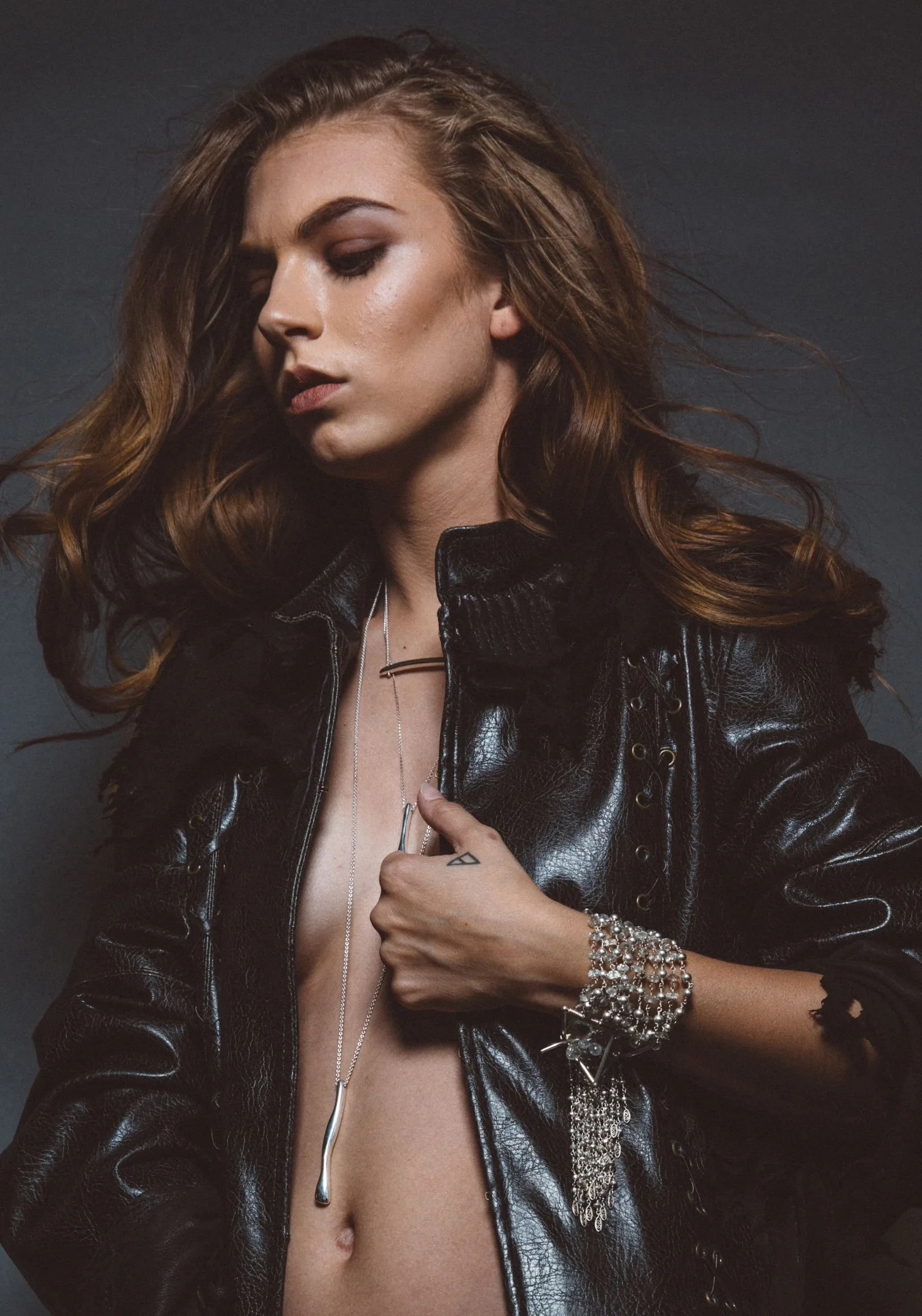 A model with long auburn hair in an open black leather jacket, wearing a long silver pendant necklace and a wide silver cuff