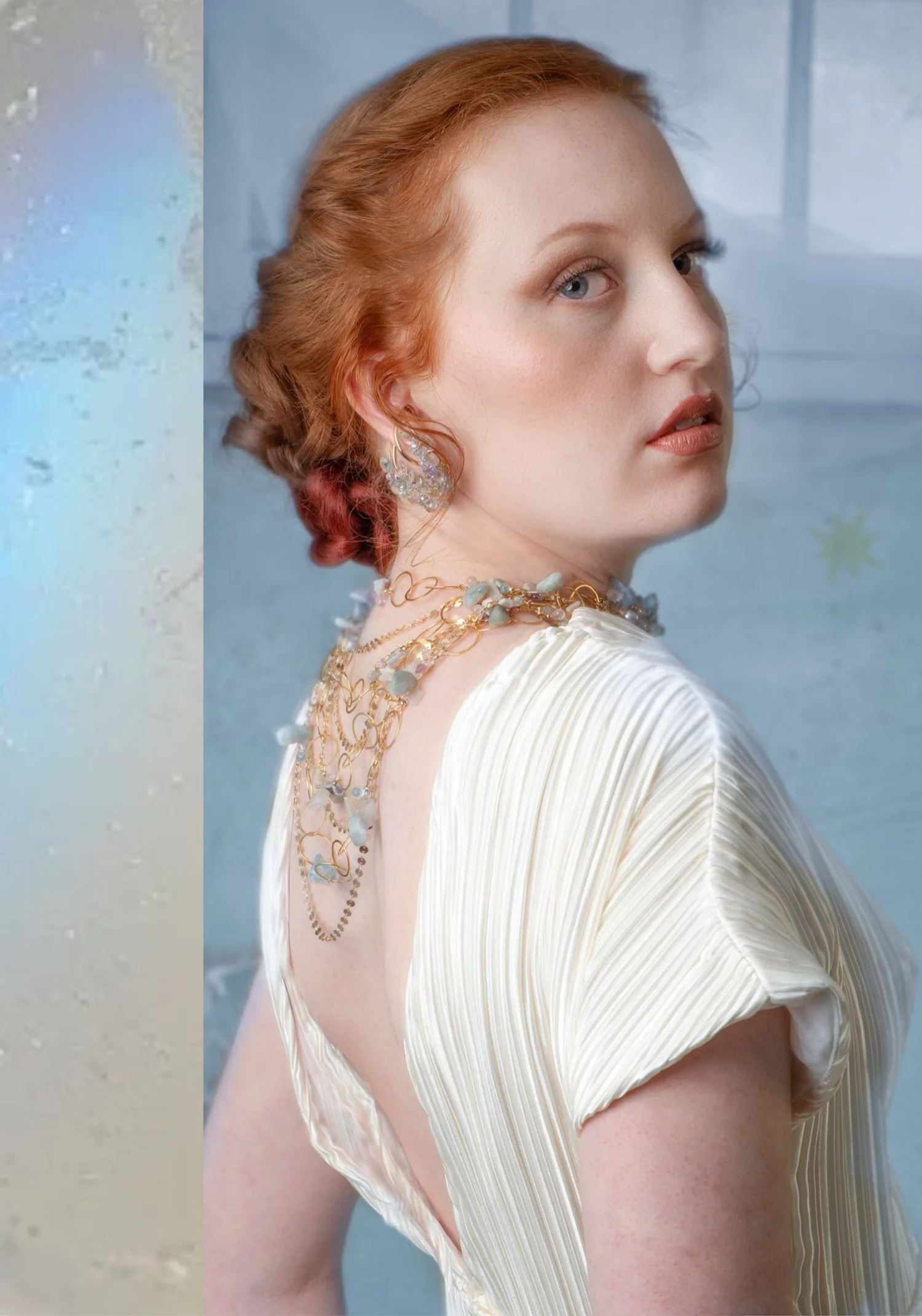 A red-haired model in a pleated ivory gown looking over her shoulder, wearing a gold ear climber and a gold wire necklace draped down her back, both clustered with pale blue gemstones