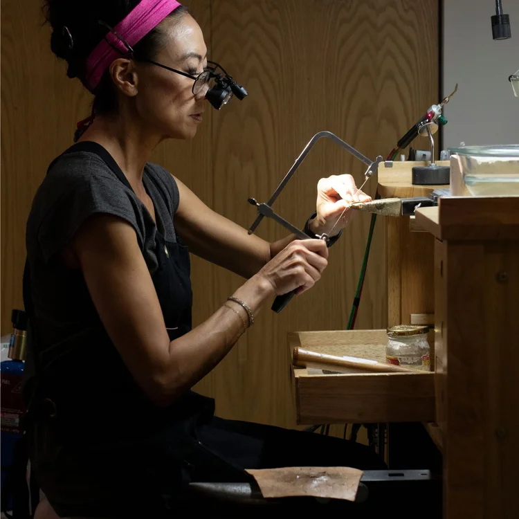 Andrea Li at her bench in a pink headband and loupes, sawing with a jeweler's saw