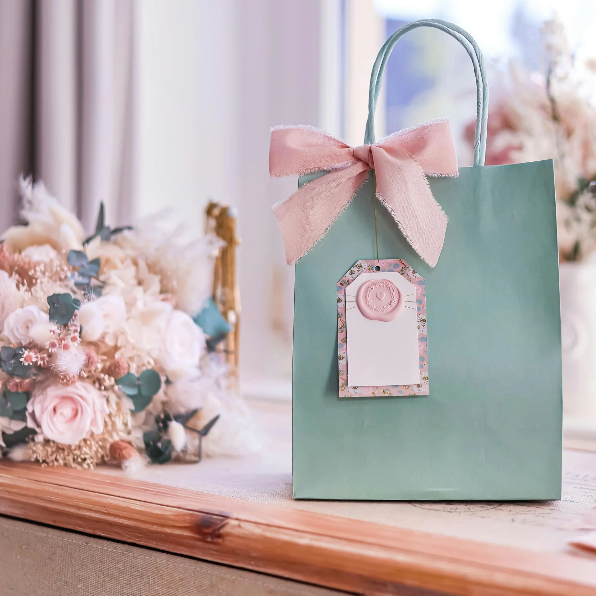 Mint green gift bag with a pink ribbon bow and decorative gift tag, displayed beside a soft blush-and-cream bouquet on a wooden surface in a bright, airy setting.