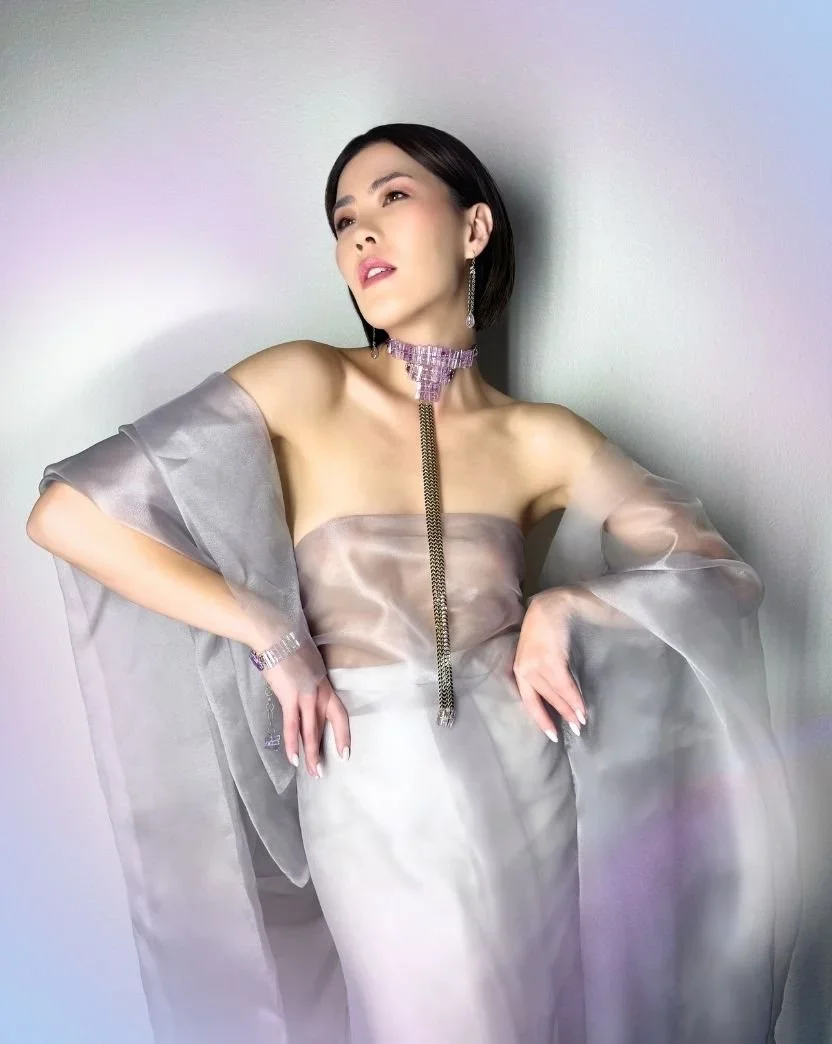 Model in a sheer off-shoulder wrap and satin dress wearing a lilac gemstone choker with a long chain drop, matching earrings, and bracelet against a soft pastel background.