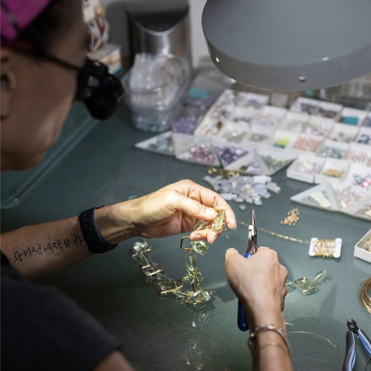 Andrea Li with pliers assembling a gold and green gemstone necklace beside trays of stones