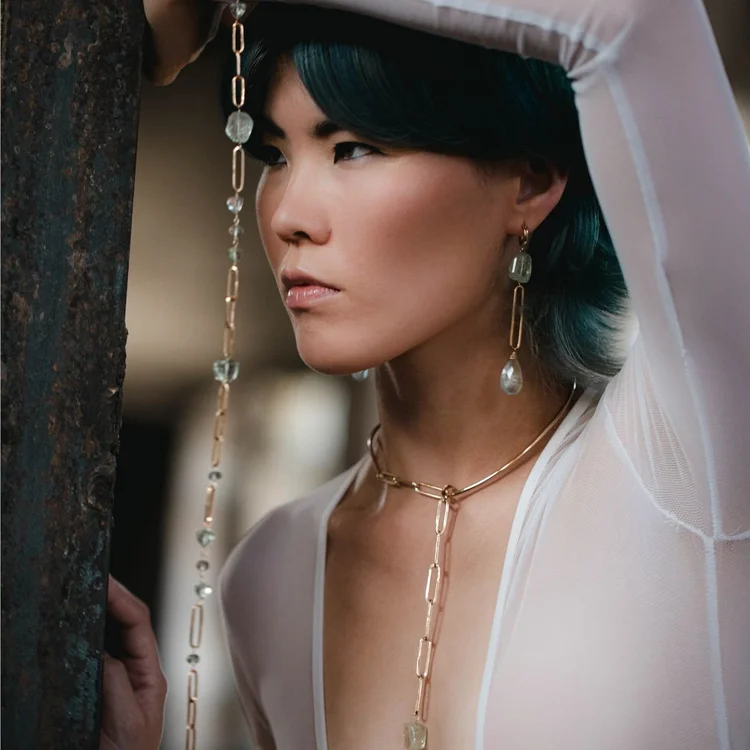 A model with teal hair in a white hooded top wearing long gold chain earrings and a lariat necklace
