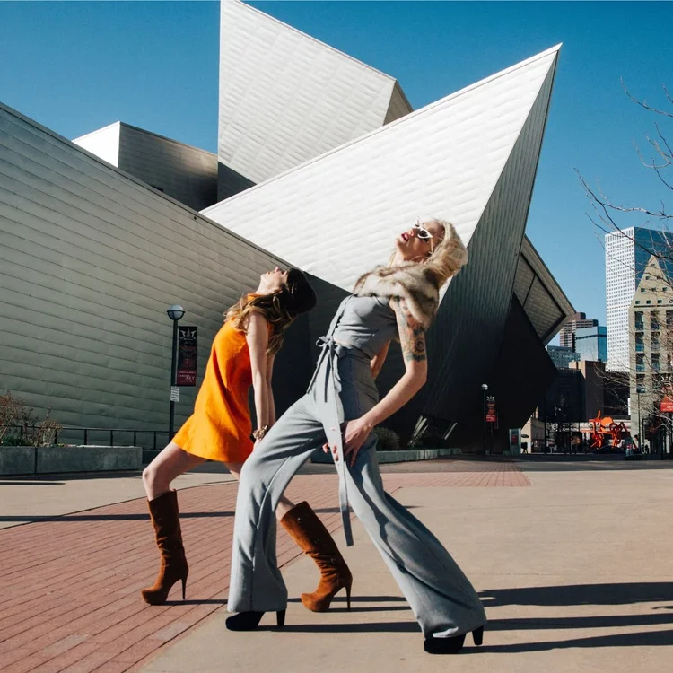 Two models in bold outfits striking poses on a plaza in front of a sharply angled modern museum building