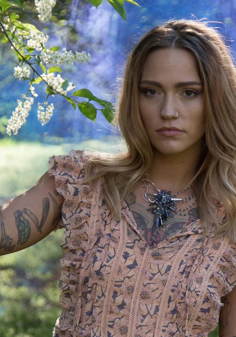 A tattooed model in a ruffled print top before blue smoke and white blossoms, wearing a dark crystal cluster pendant