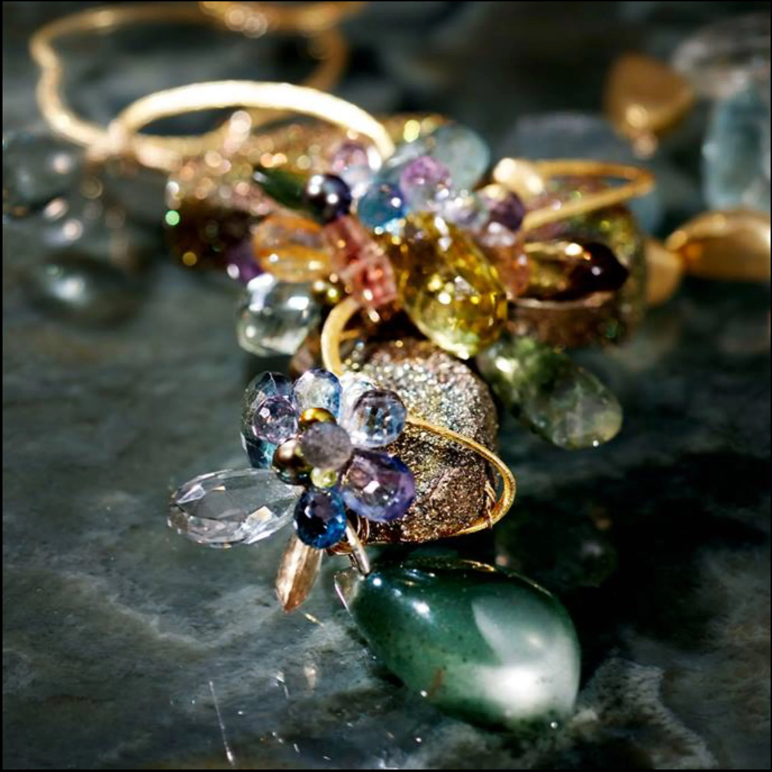 Close-up of colorful gemstone rings on a dark glossy surface, featuring floral bead clusters, gold bands, and sparkling crystals.