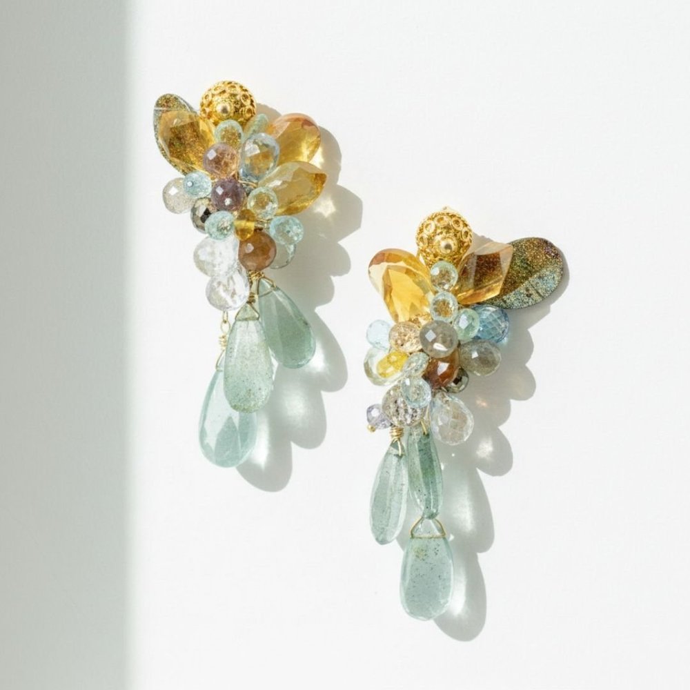Close-up of pastel gemstone earrings with golden accents and pale aqua drops, casting soft shadows on a white background.