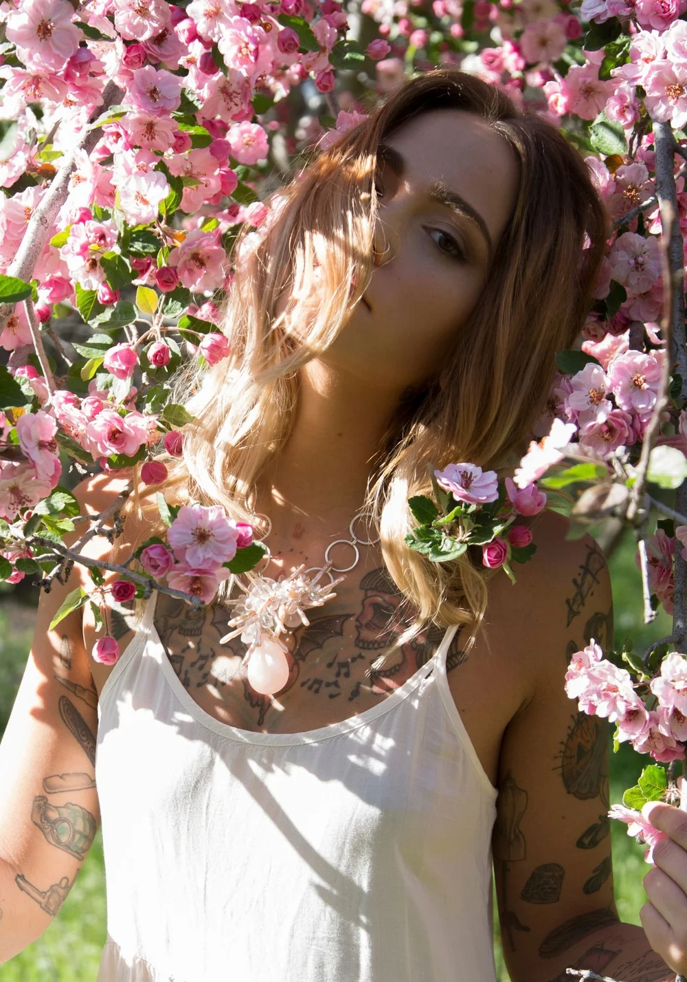 A tattooed model in a white camisole among pink blossoms, wearing a pale crystal statement necklace