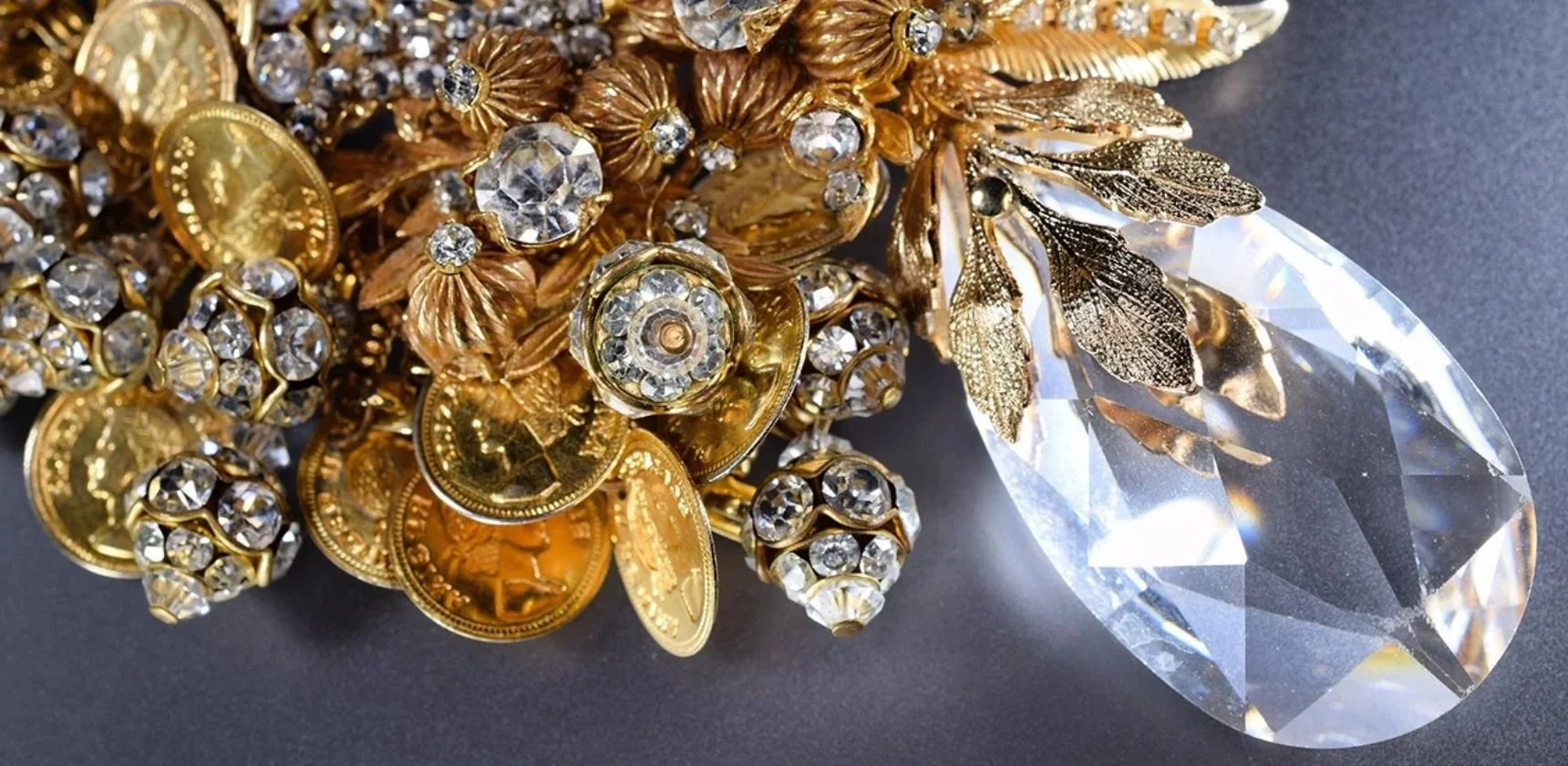 Vintage gold jewelry close up: clusters of gold coins, rhinestone beads and gold leaves around a large clear crystal teardrop