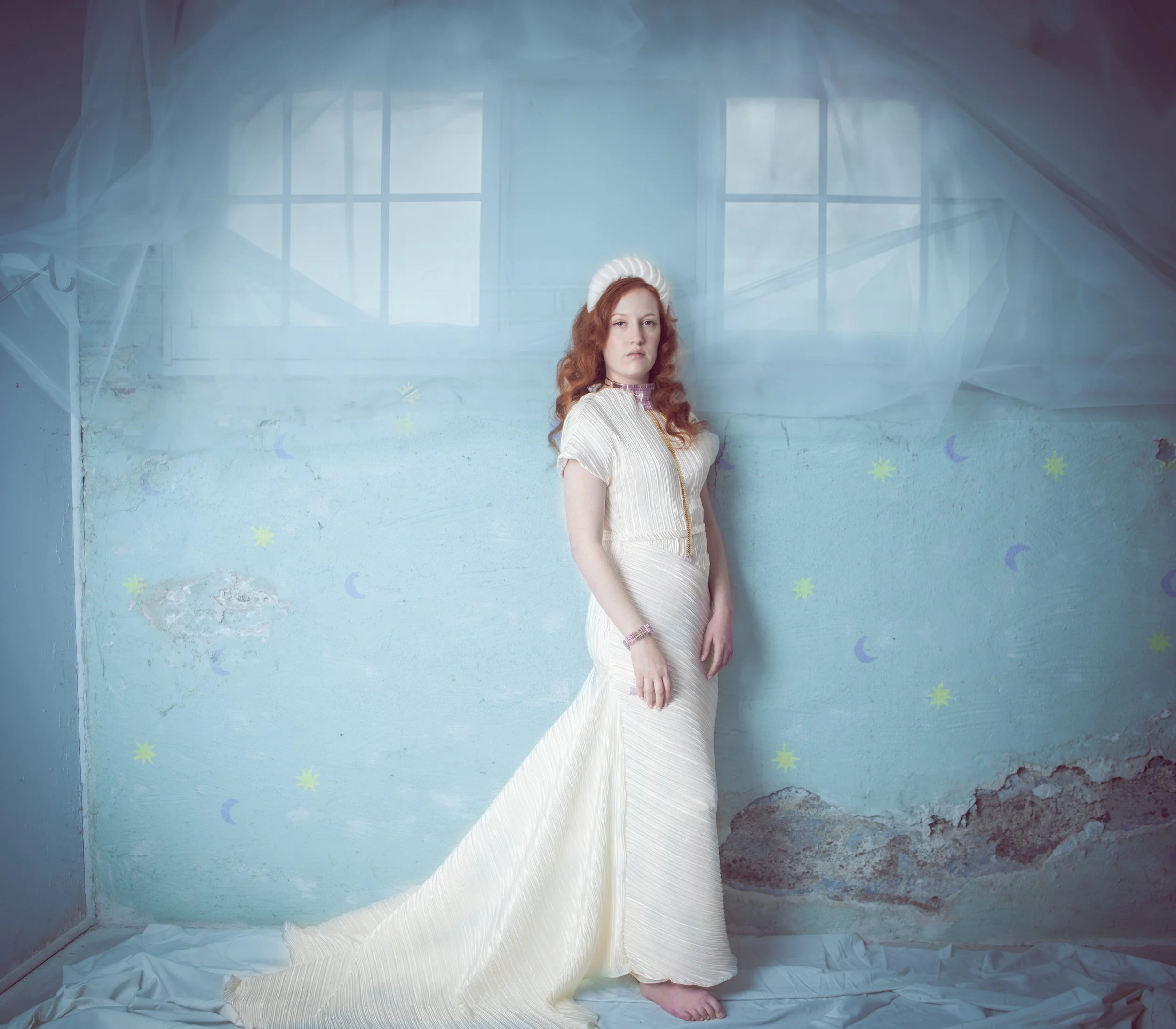 Full-length portrait of a red-haired woman in a flowing white gown, standing barefoot against a dreamy pale blue backdrop with sheer drapery.