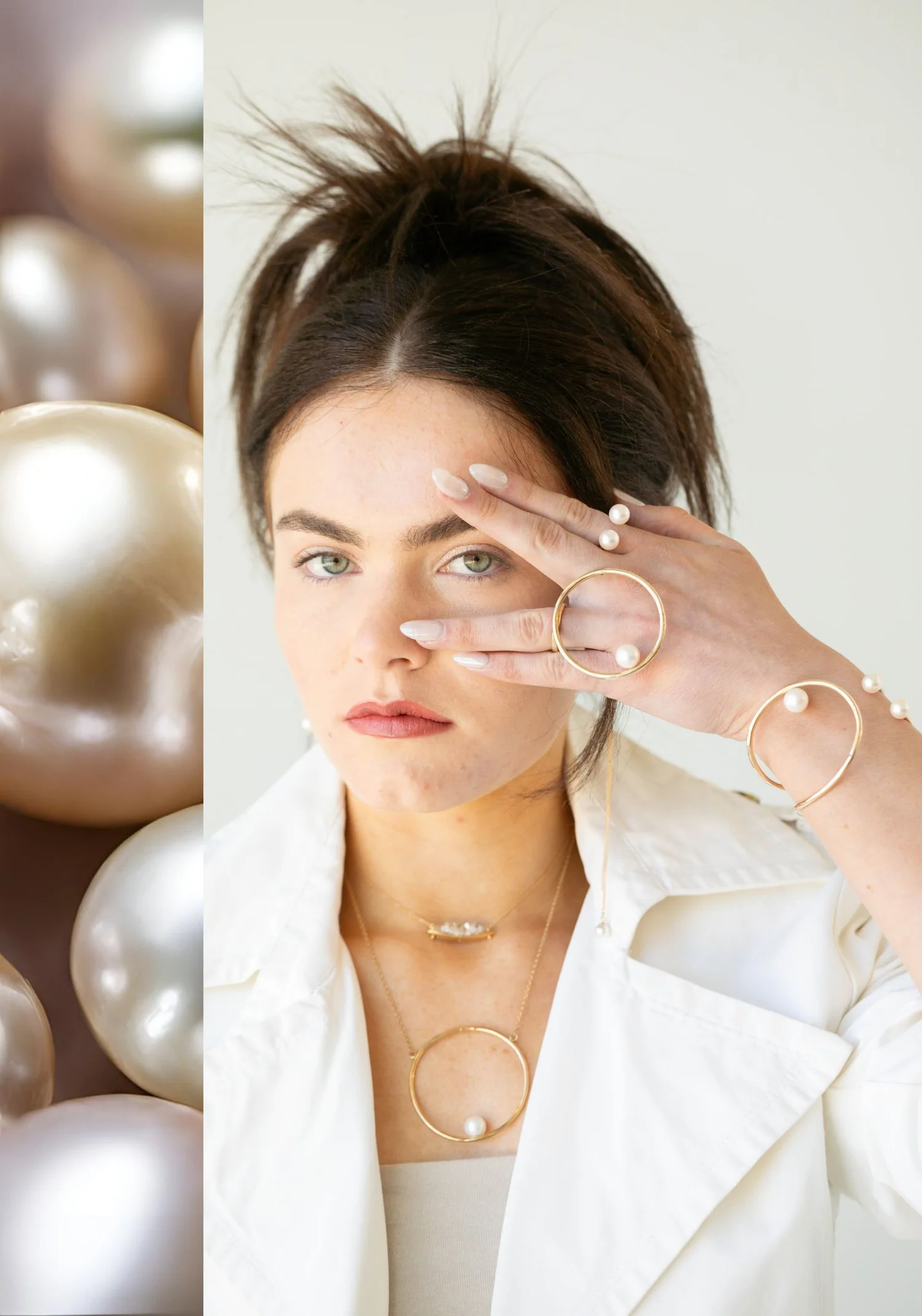 A model with dark hair in a white jacket, one hand raised across her face wearing a gold open circle pearl ring, with a gold pearl bangle and a gold circle pendant with a pearl, beside a close-up of large lustrous pearls