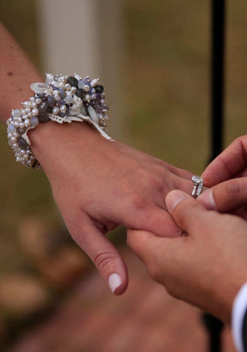 Close-up of a hand wearing a custom bridal wrist cuff made of pearls and lavender-gray beads while another hand gently places a ring on the finger.