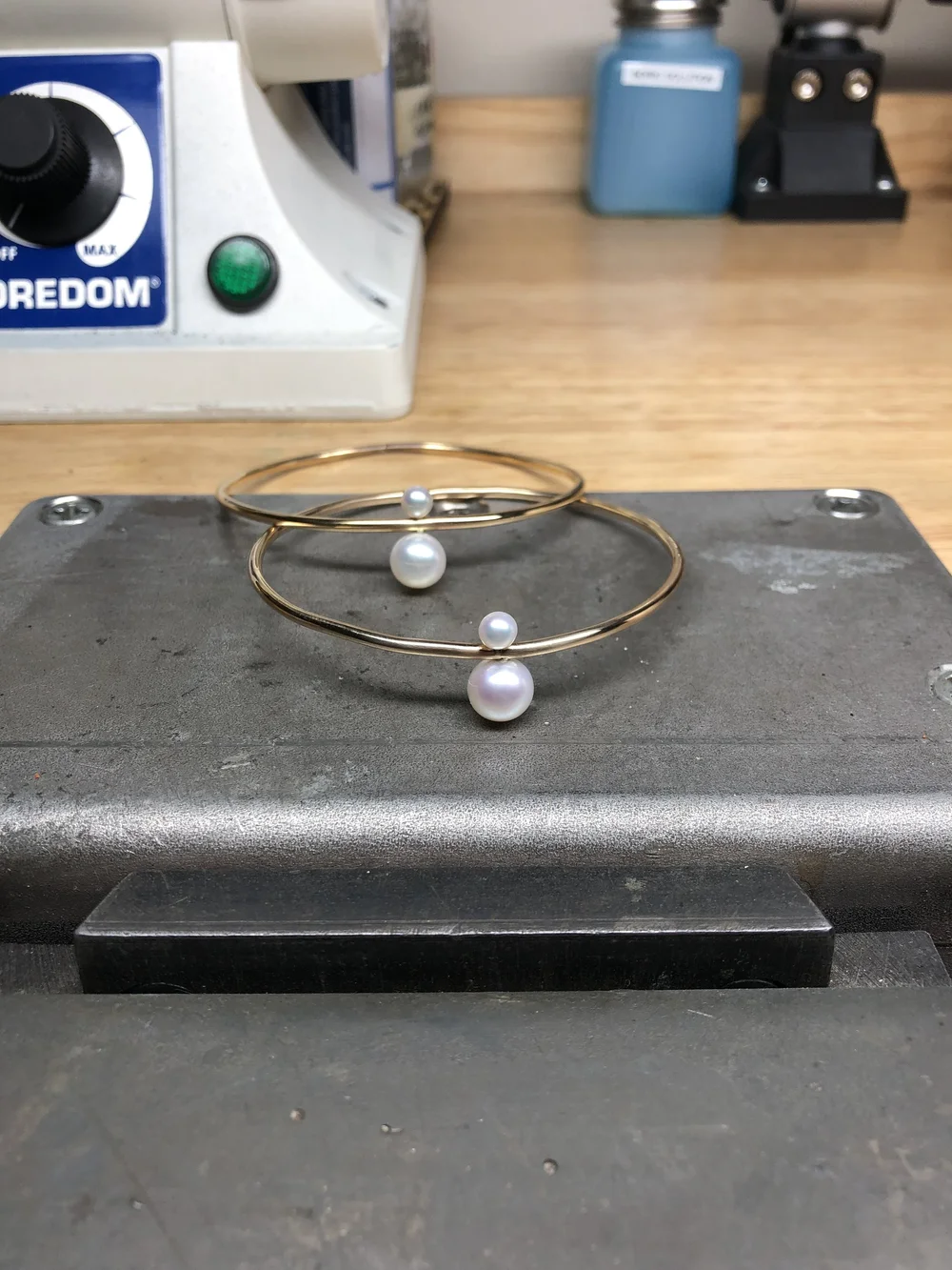 Two gold wire bangles set with white pearls resting on a steel bench block at Andrea Li's bench, a Foredom flex shaft motor behind them