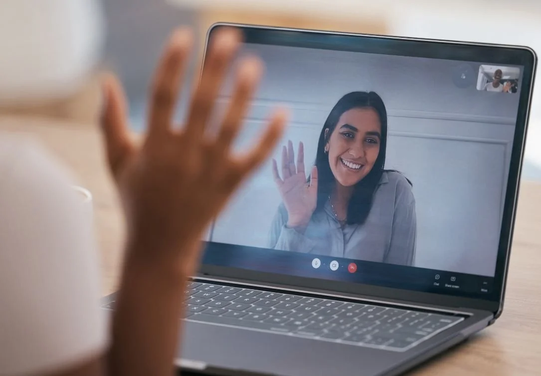 Laptop screen showing a smiling woman waving during a video call, with a blurred hand in the foreground.