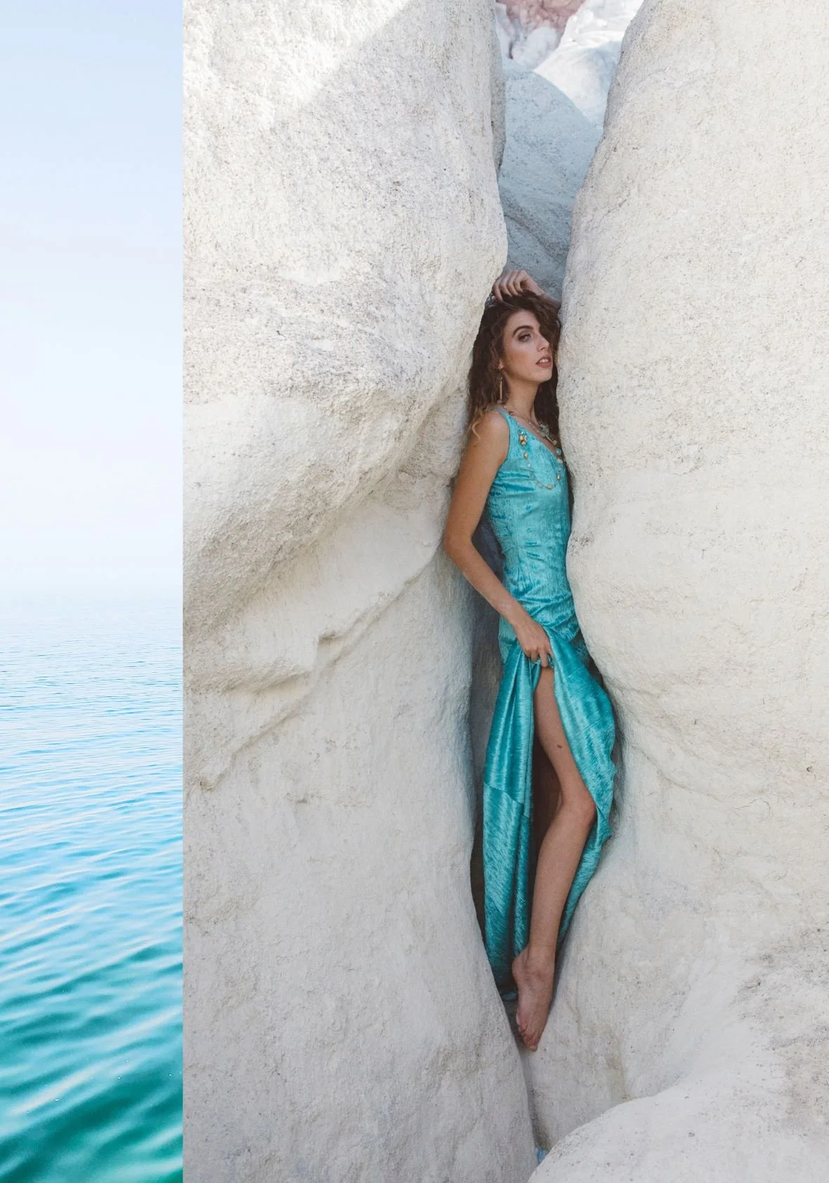 Woman in a shimmering turquoise dress stands between white rock walls, with a narrow strip of calm blue water along the left edge.