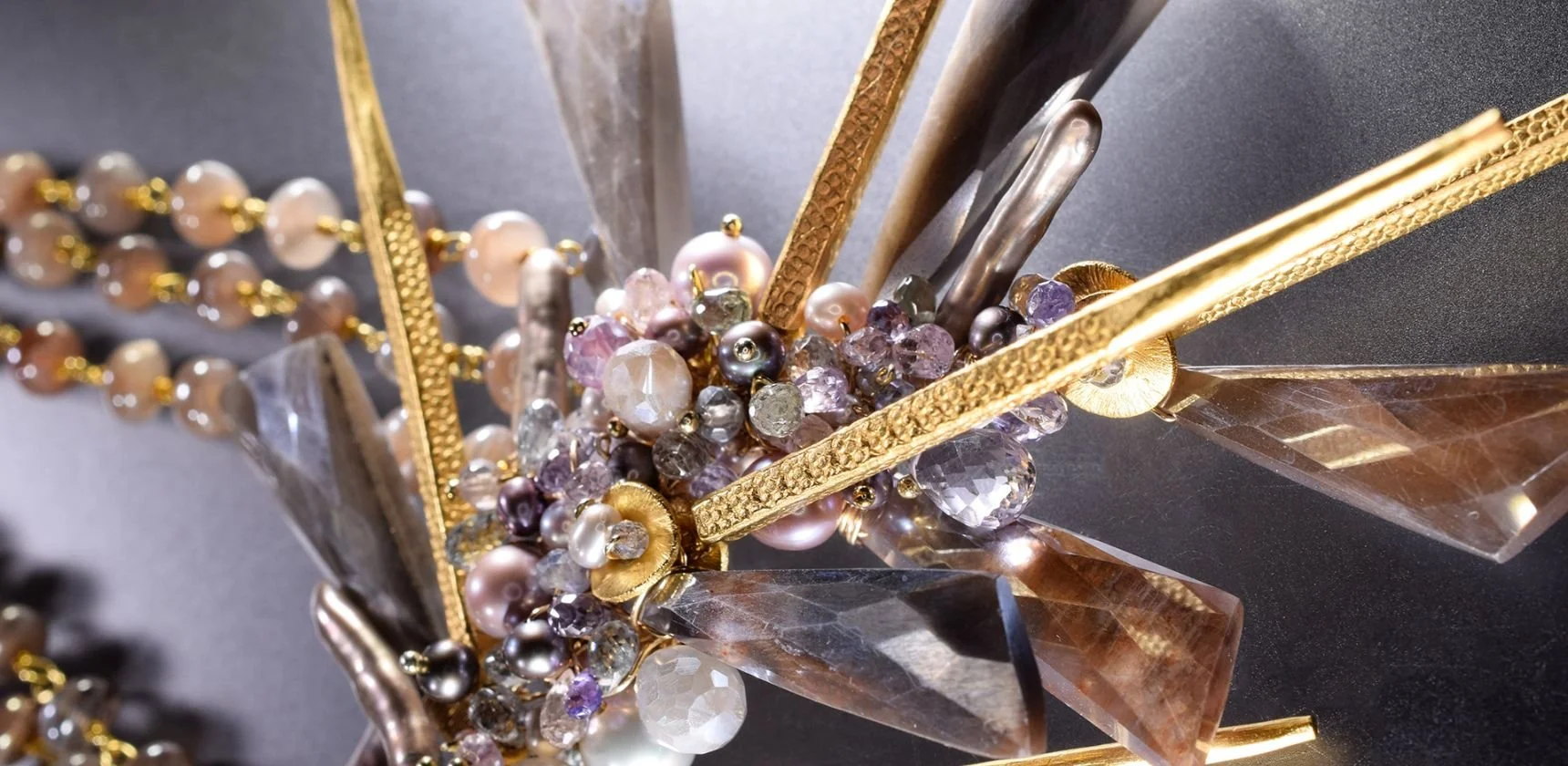 Close view of an Andrea Li statement necklace: hammered gold spikes and faceted crystal points around a cluster of pearls and pastel gemstones, beside a strand of peach moonstone beads