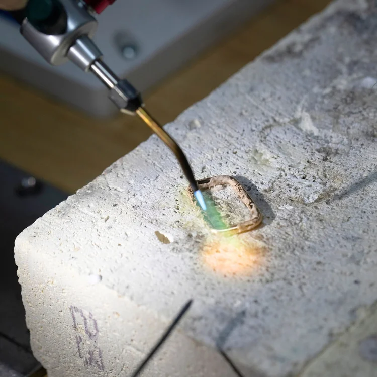 A torch flame soldering a small gold frame around a pale green stone on a soldering block
