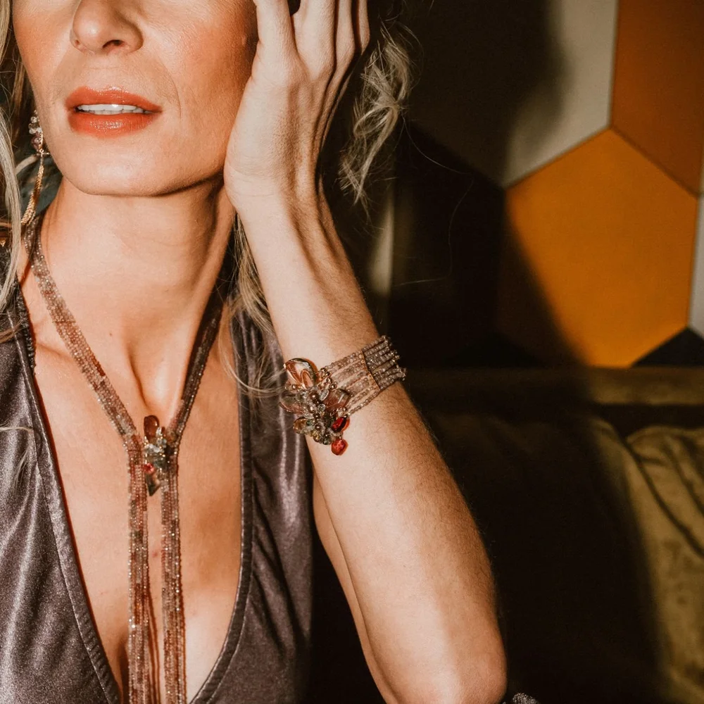 A model in a bronze satin top touching her face, wearing an Andrea Li gemstone cluster bracelet and a long beaded lariat