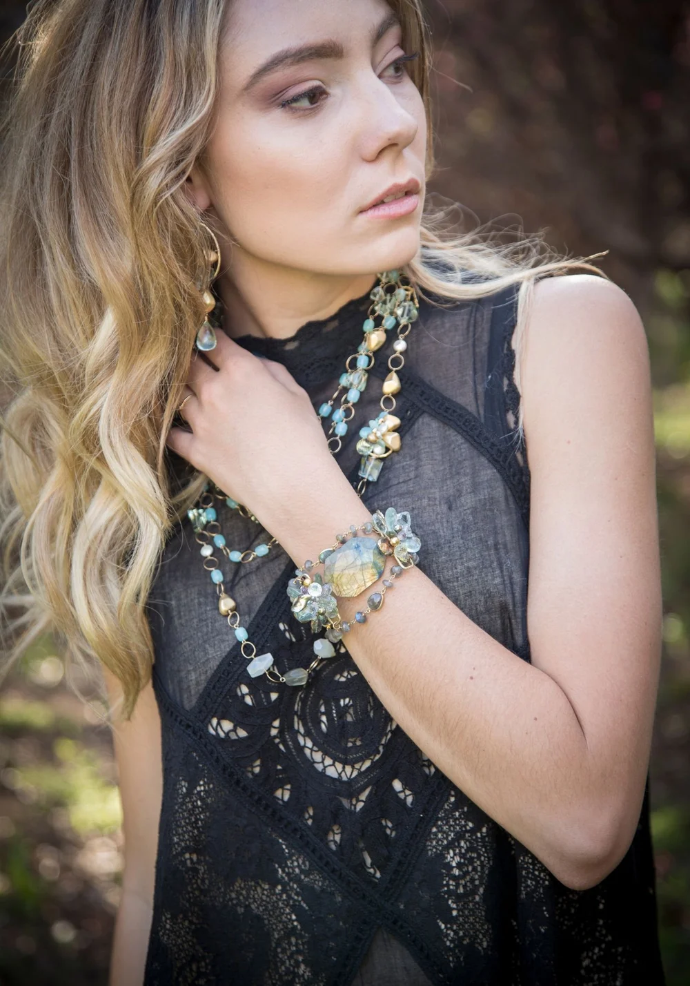 A model in black lace looking over her shoulder, wearing long gemstone necklaces and a stacked gemstone bracelet