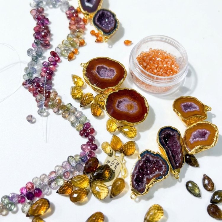 Gold-edged geode slices with purple crystal centers, strands of multicolor faceted sapphire beads, honey-colored faceted teardrops and a jar of small orange stones on a white table