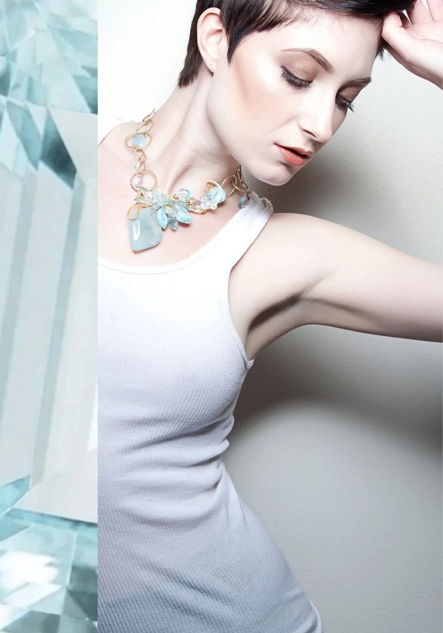 A model with short dark hair in a white tank top wearing a gold link necklace set with aquamarine stones and a large aquamarine drop, beside a close-up of faceted aquamarine