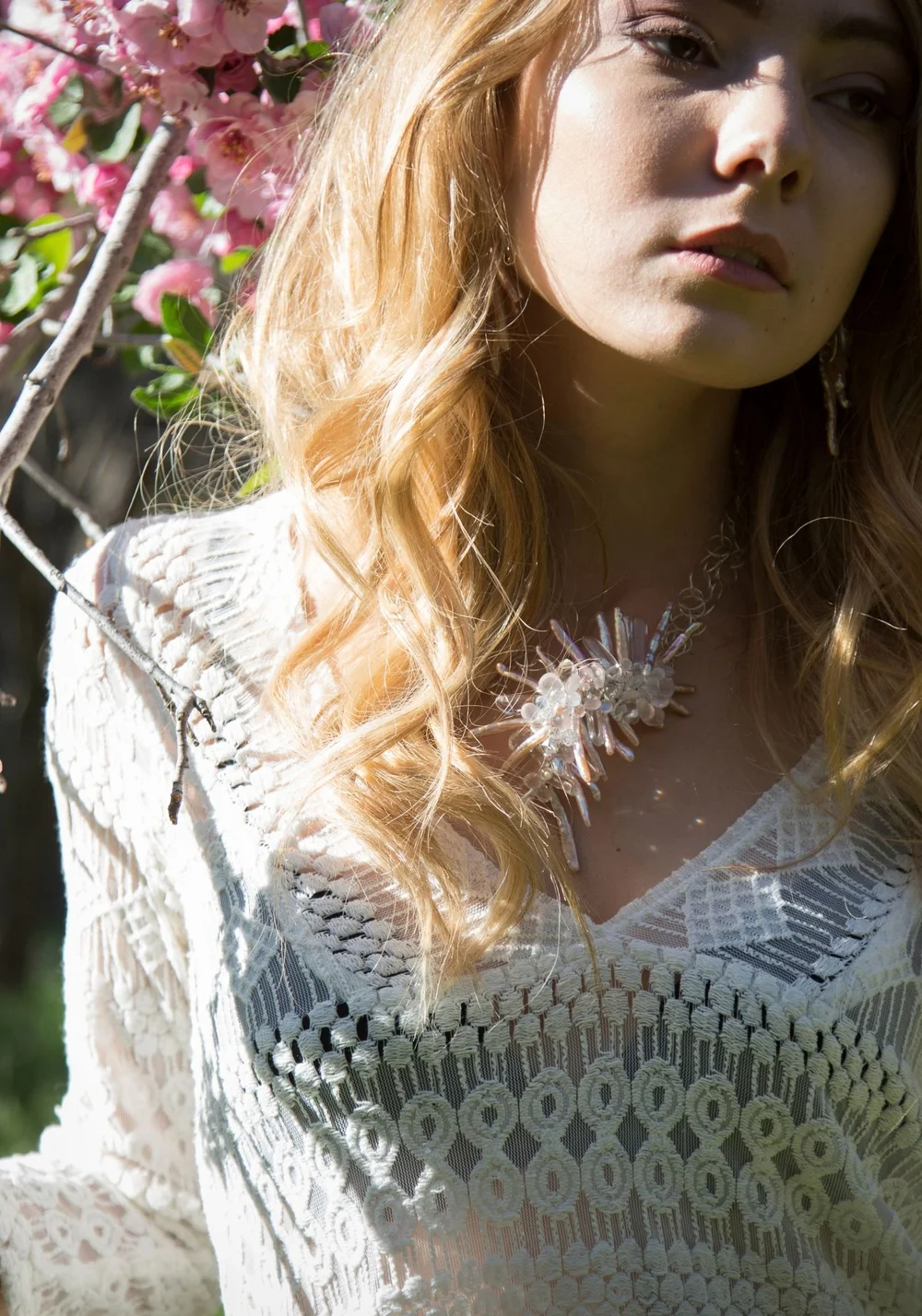 A model in white crochet lace in soft light among pink blossoms, wearing a pale crystal cluster necklace