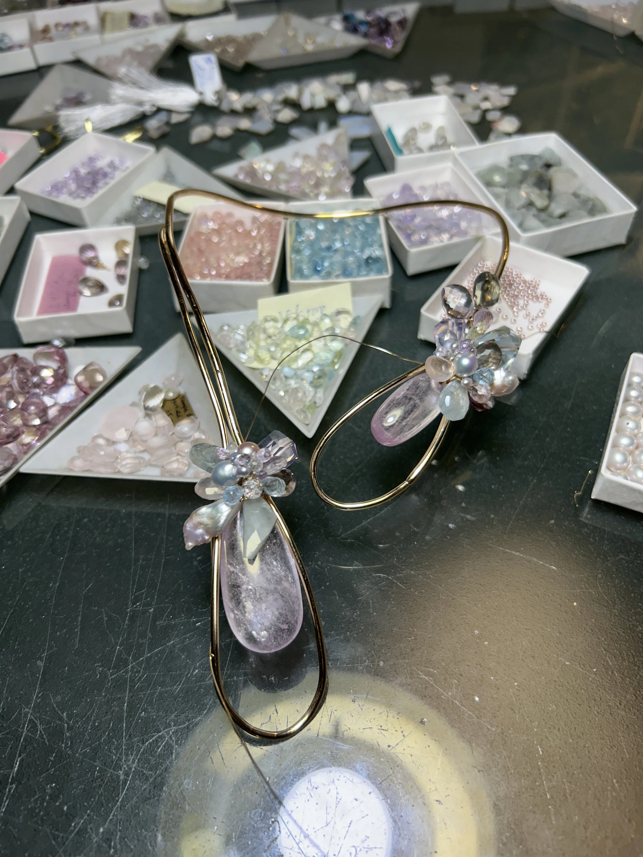 Sculptural gold wire choker in progress, each with a pale pink teardrop gemstone and a clustered mix of pastel stones and pearls, photographed on a jewelry workbench surrounded by trays of loose gemstones.