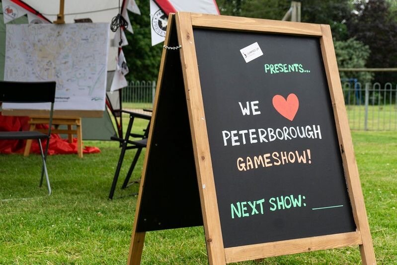 Chalk board on grass with writing on it that says ‘Jumped Up Theatre presents… We love Peterborough Gameshow! Next show: (blank)’