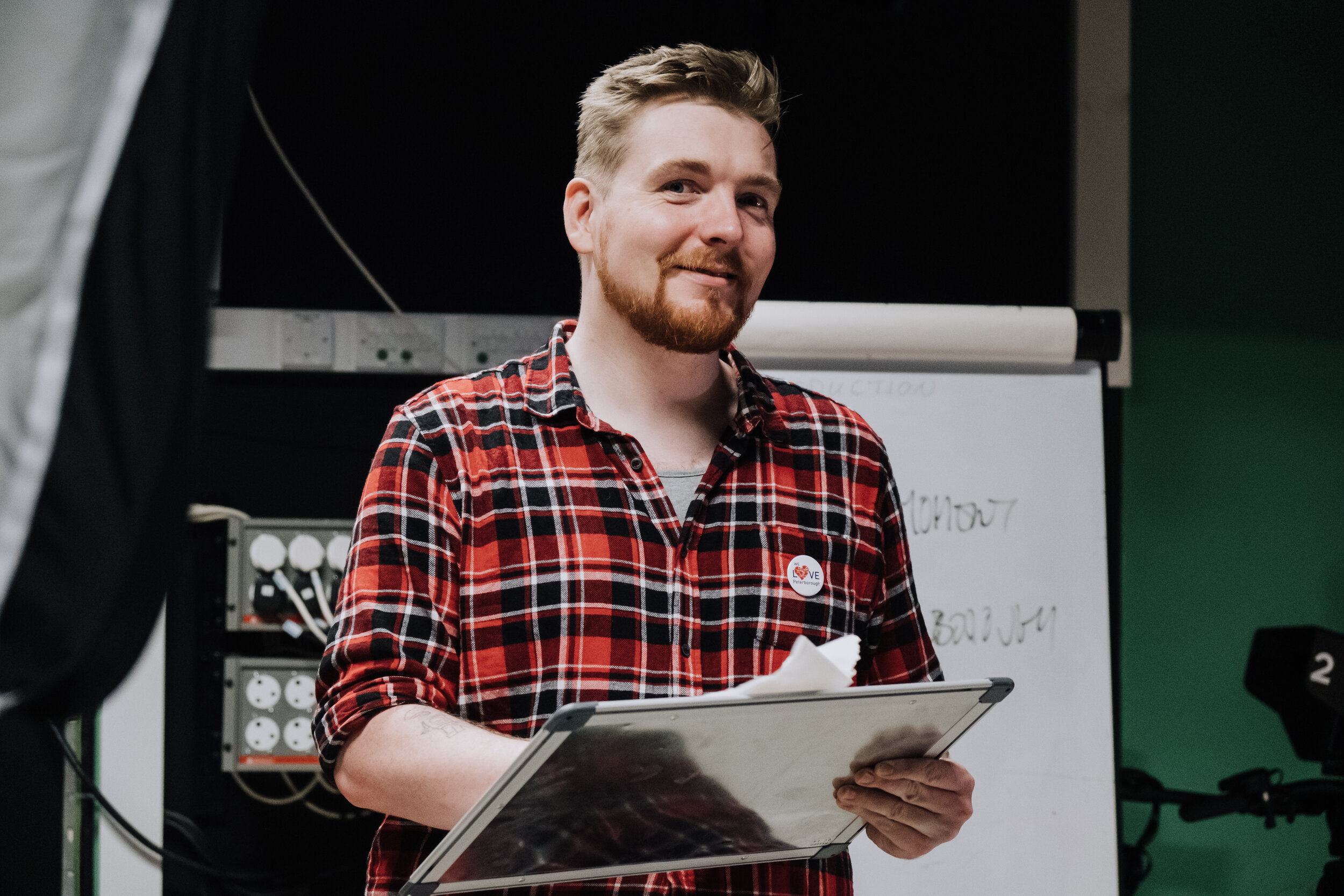 A image of a white man with reddish hair in a red plaid top stnading at what looks like a music stand. He has just caught sight of the camera, is looking directly at it, and smiling.