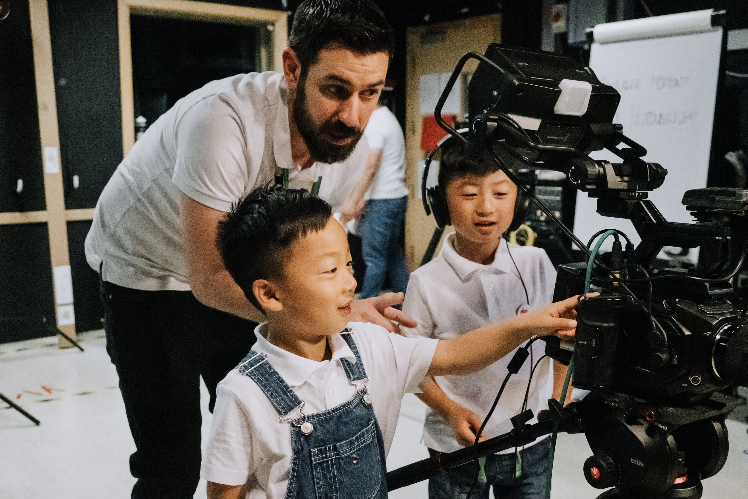One adult man with dark hair and a slight beard is teaching two children how to operate a professional camera. One of the children is wearing over-ear headphones while they do this.