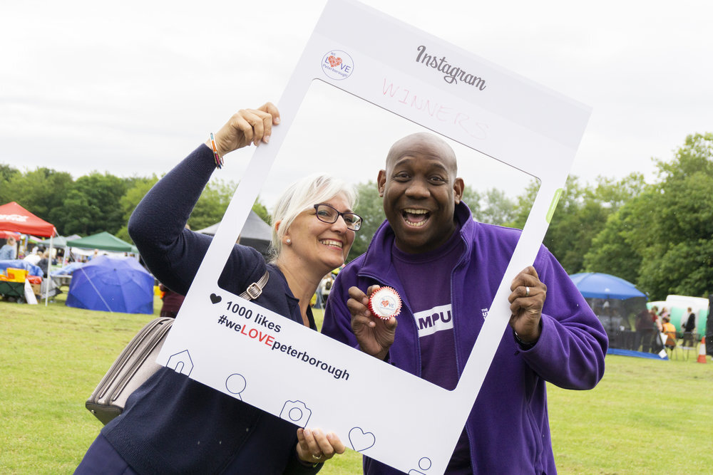 A white woman and a black man, holding up an instagram style card board frame around their faces. Both of them are smiling. The frame reads #WeLovePeterborough’ across the bottom.