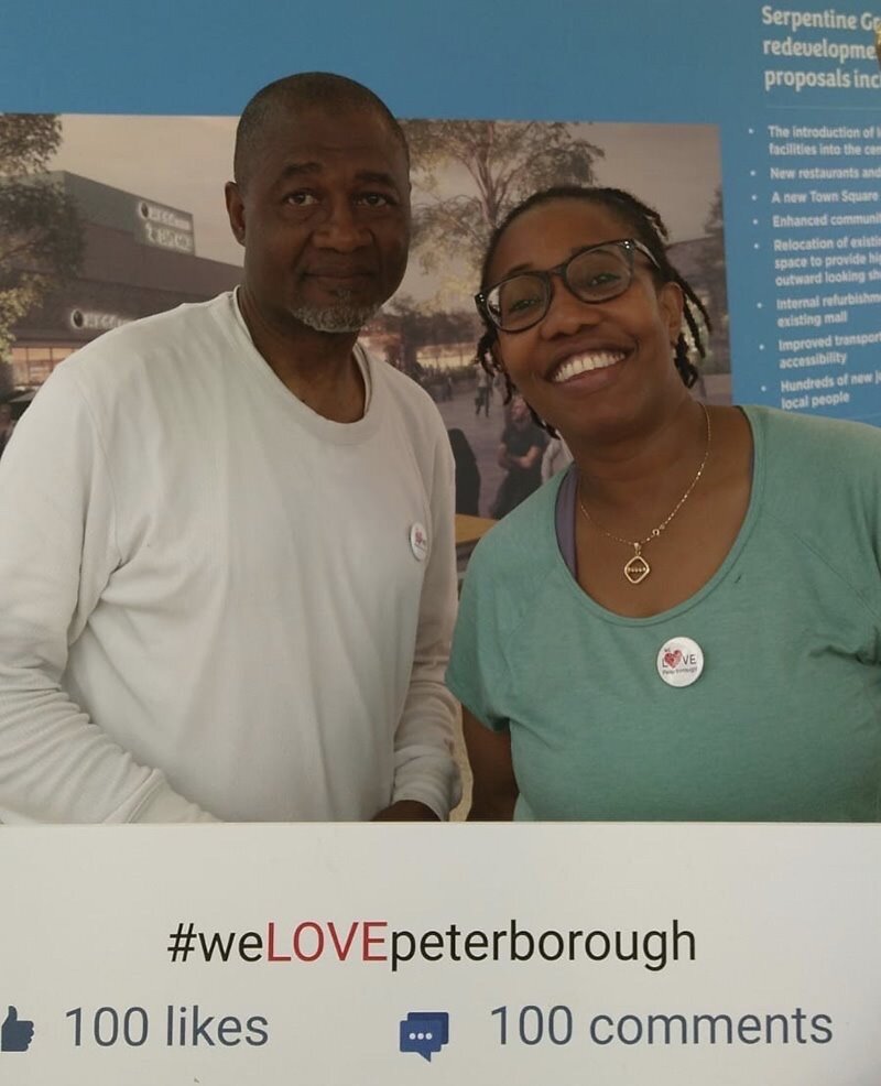 Image of two people, a black man and a black woman. They are both smiling. The man is wearing a white top and the woman glasses and a blue top with a We Love Peterborough sticker on it. 