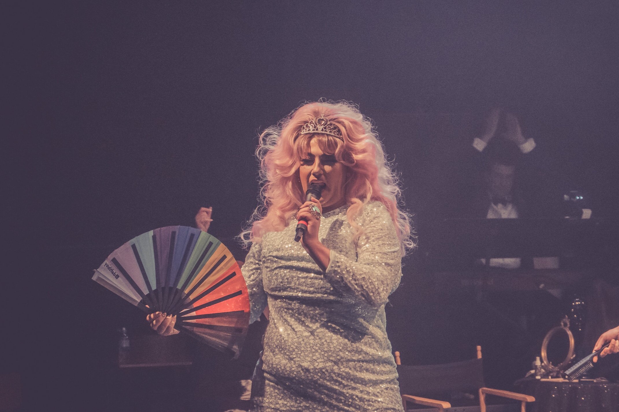 Image of a person in a pink wig, tiara and silver dress holding a rainbow fan singing into a microphone.