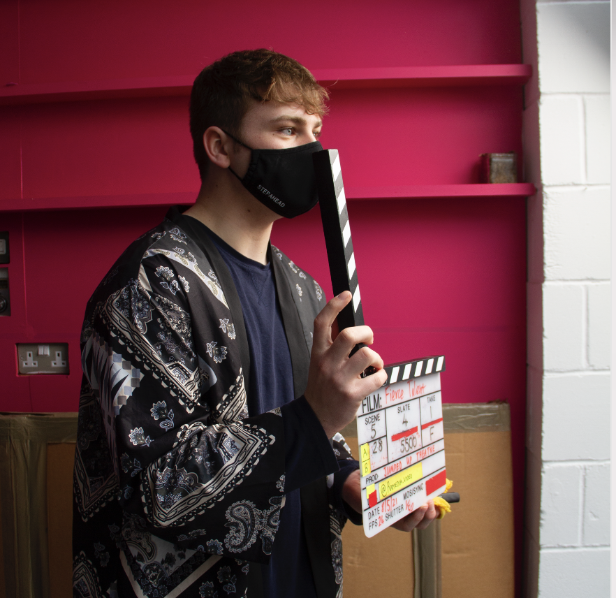 Young man with white skin and brown hair. He is holding a clapperboard and wearing a black face mask.