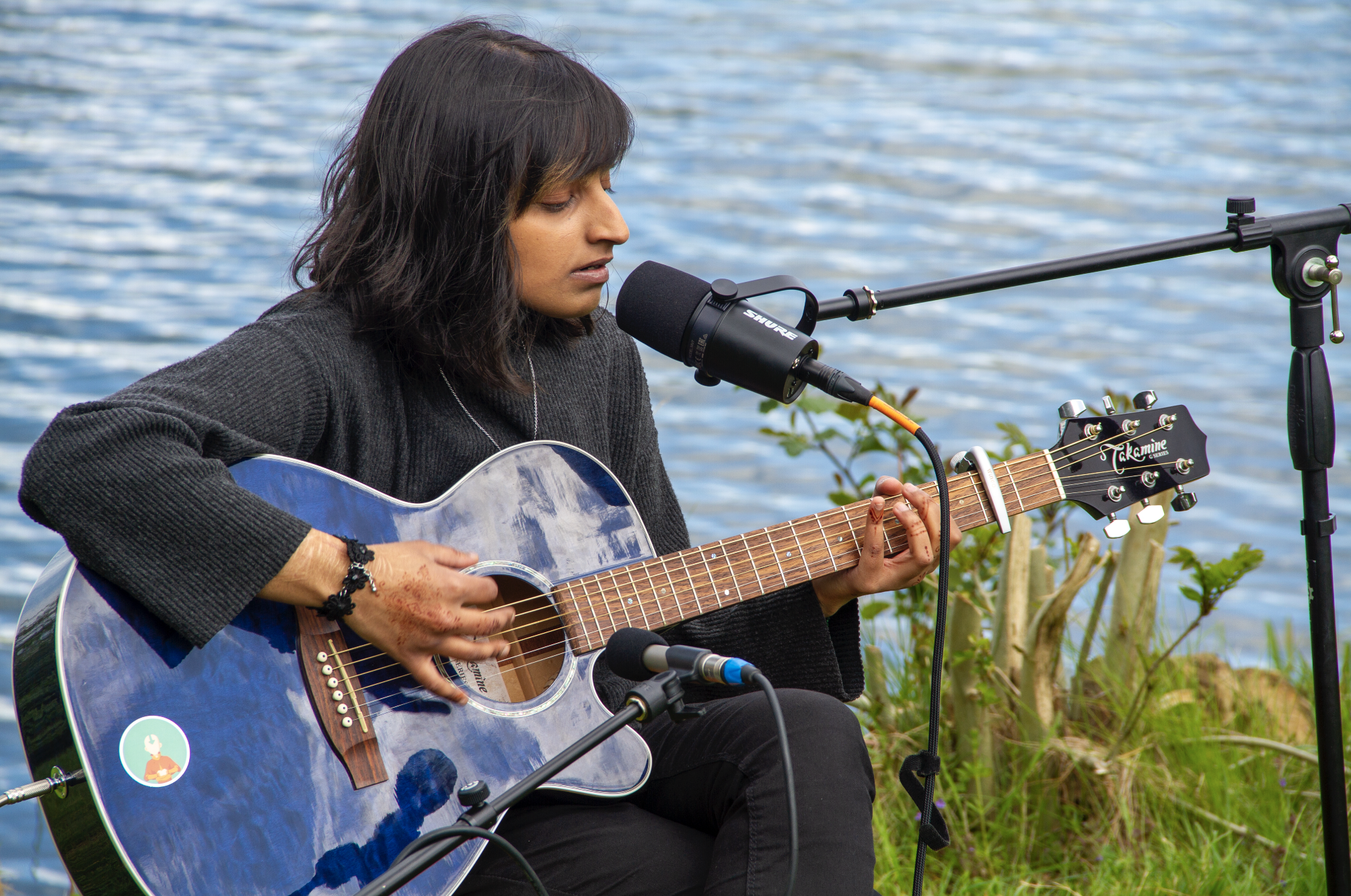Young woman singing into a microphone, she is playing a blue guitar. She has black hair and brown skin.