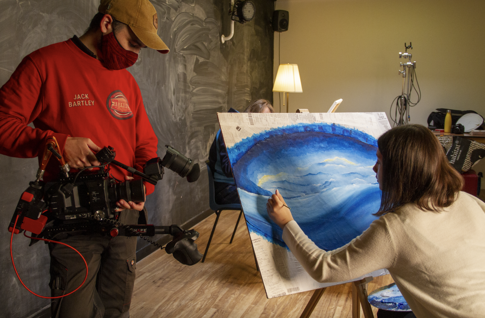 a filmmaker wearing red uses a large camera to record a female painter who is painting blue onto a large canvas