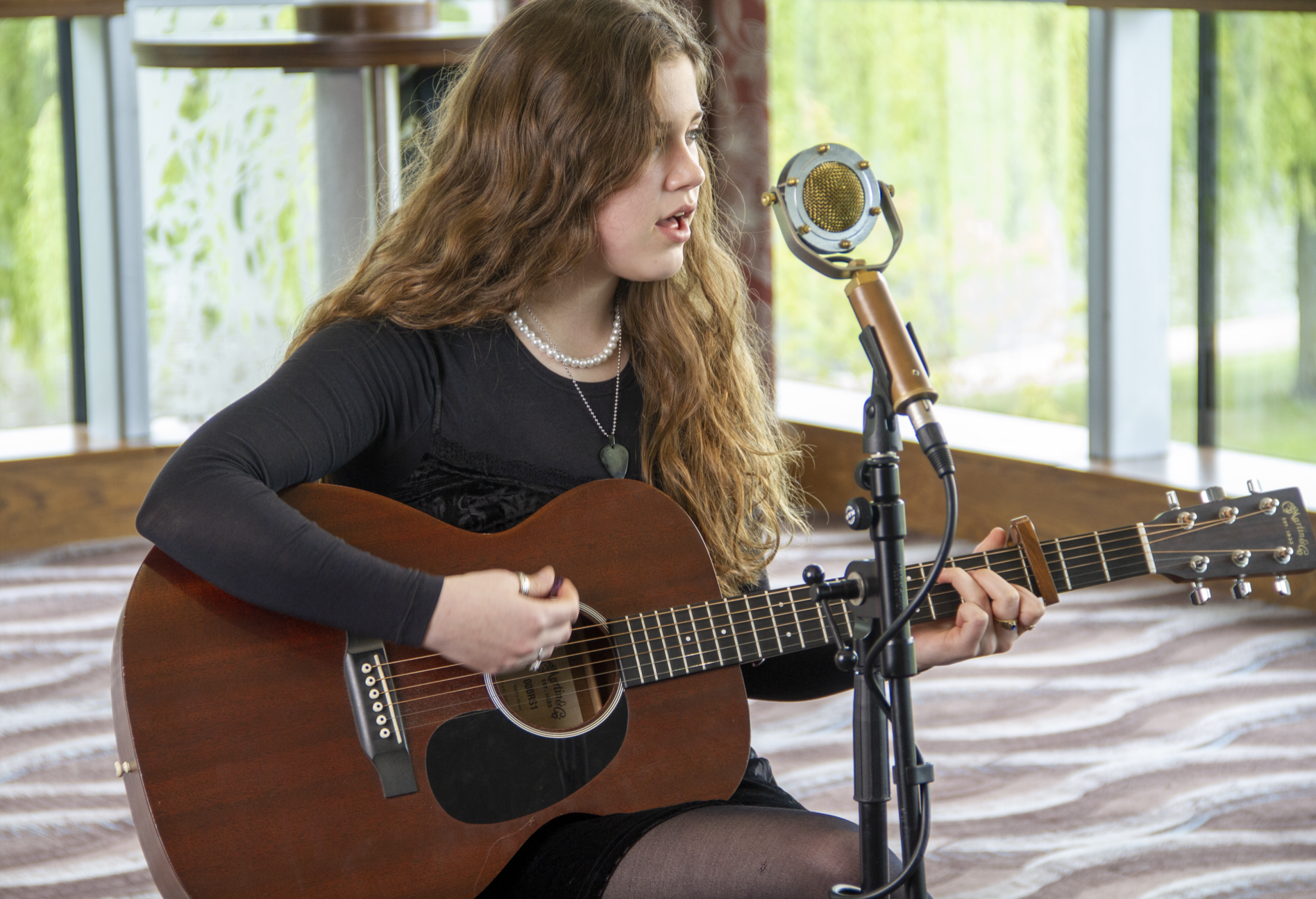 a young woman with white skin and brown hair sings into a microphone, she is playing a guitar and wearing all black.