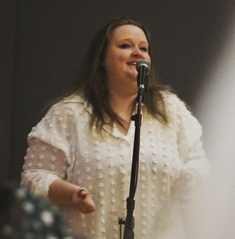  image of poet Kat Beaton, a white woman in a white shirt with brown hair, stood at a microphone delivering her poetry 