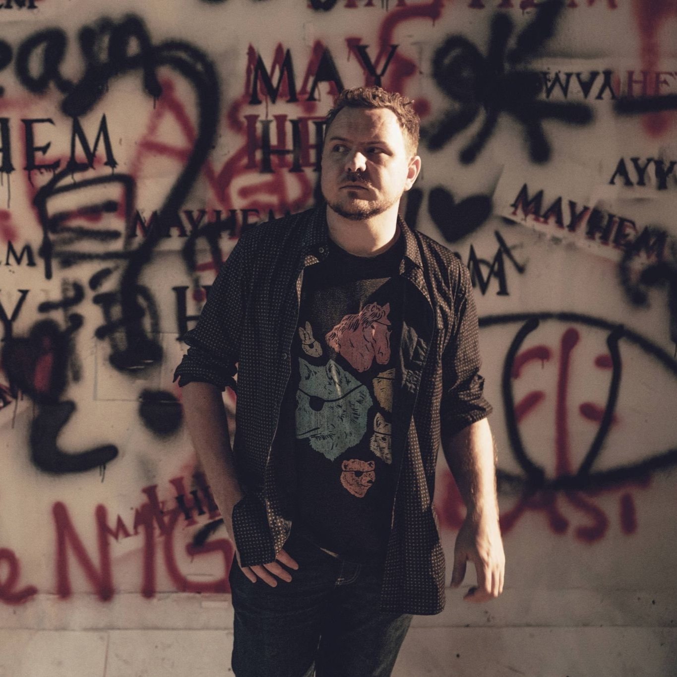  image of poet Mark Grist, a white man wearing an open brown shirt with a top underneath and jeans, posed walking away from a graffiti laden wall 