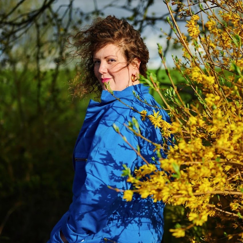  image of poet Talia Randall, a white woman with short brown hair blowing in the wind, wearing a blue trench coat posed outside next to yellow foliage that is also windswept 