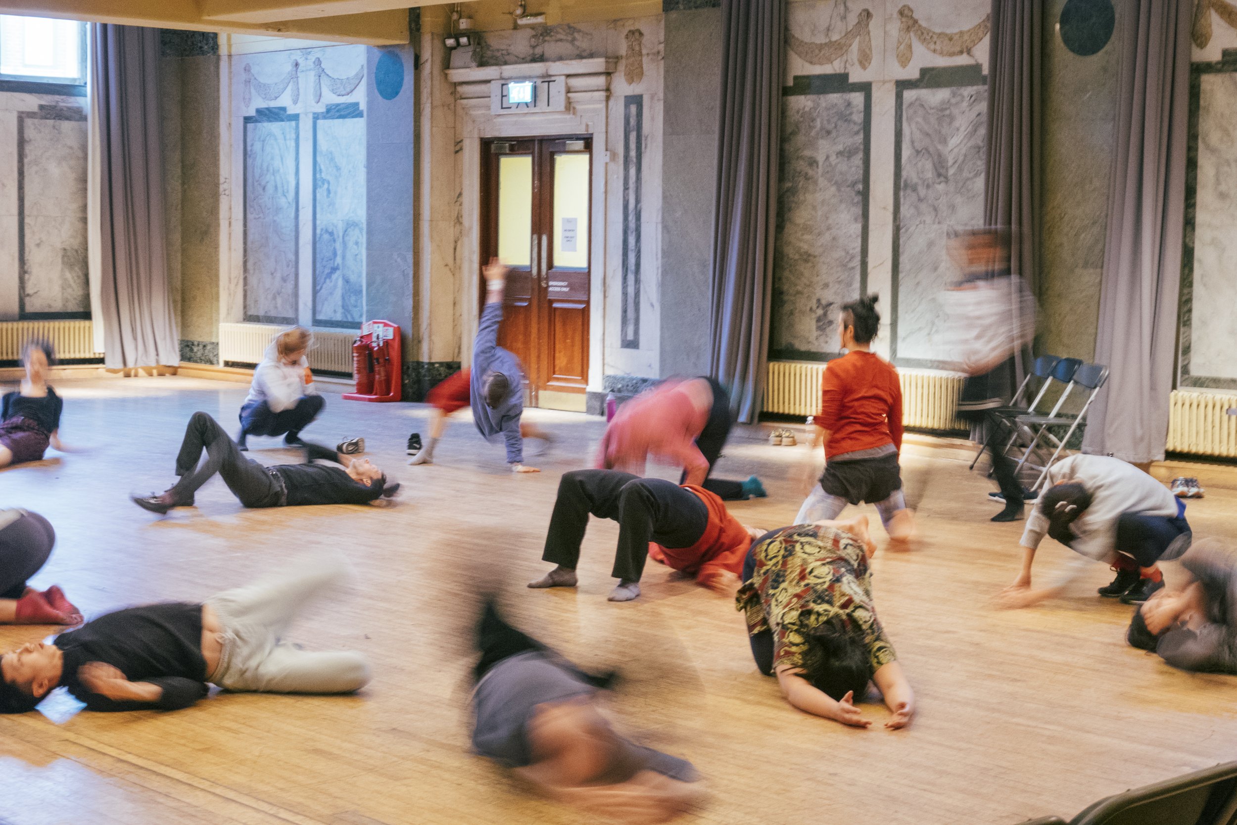  image of a wooden floored dance hall full of blurred people in colourful exercise clothes, all mid dance, stretch, or movement 