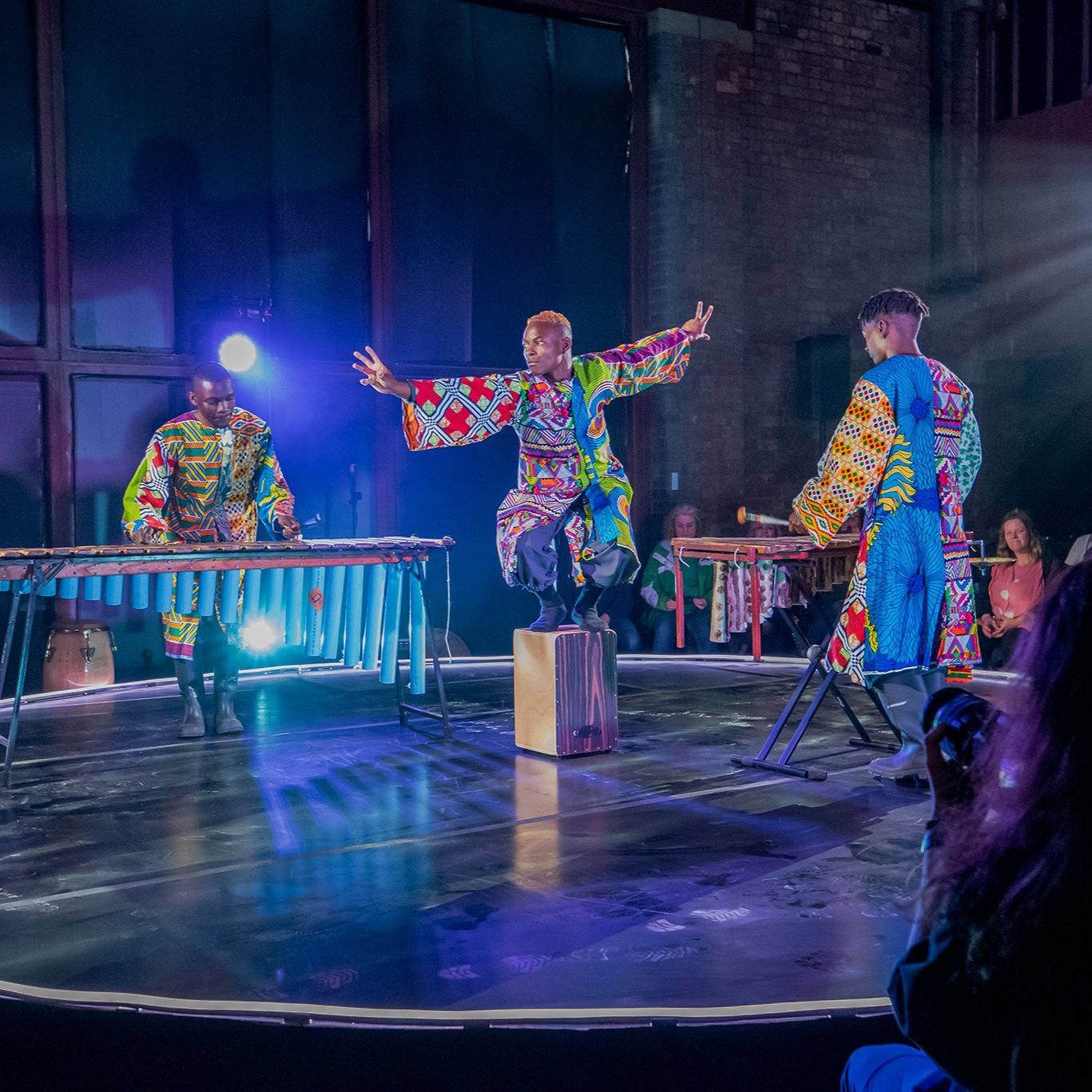  Three Black men in colourful clothing inspired by traditional African dress on a blue-lit stage 