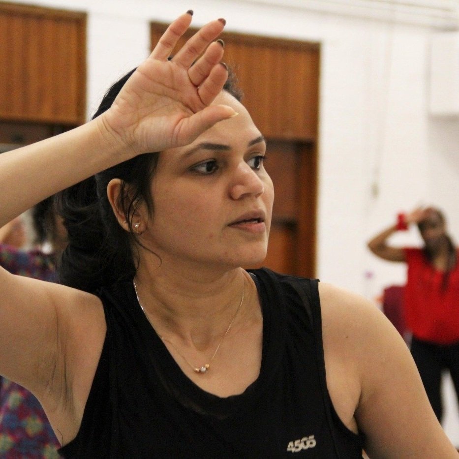  A South Asian woman with dark hair in a ponytail in a dance class. Her hand is up to wipe her forehead 
