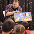 An adult male reading a children's book to young children.