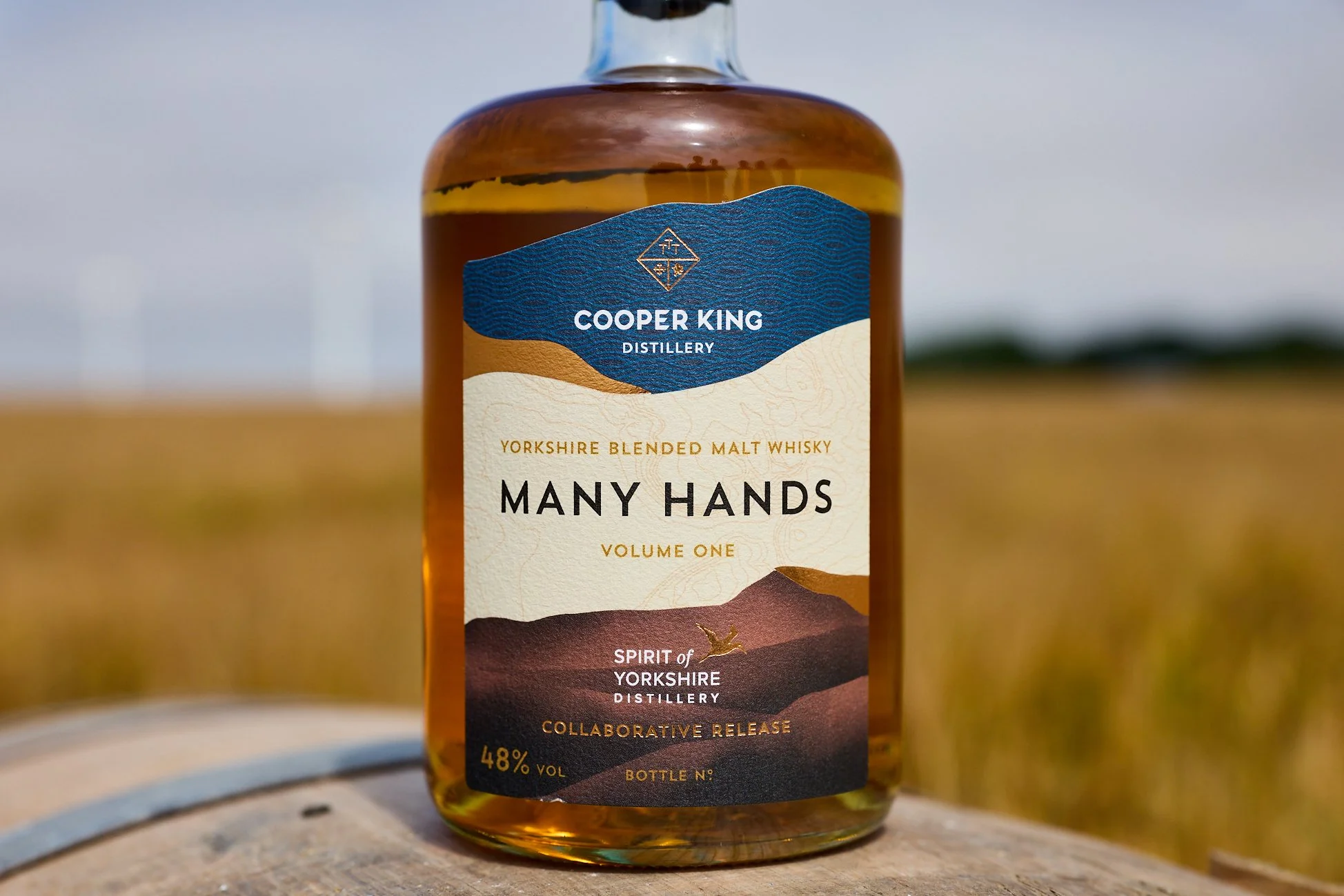 A bottle of Many Hands whisky, balanced on a cask, in front of a field of barley in the Yorkshire Wolds, with wind turbines on the horizon.