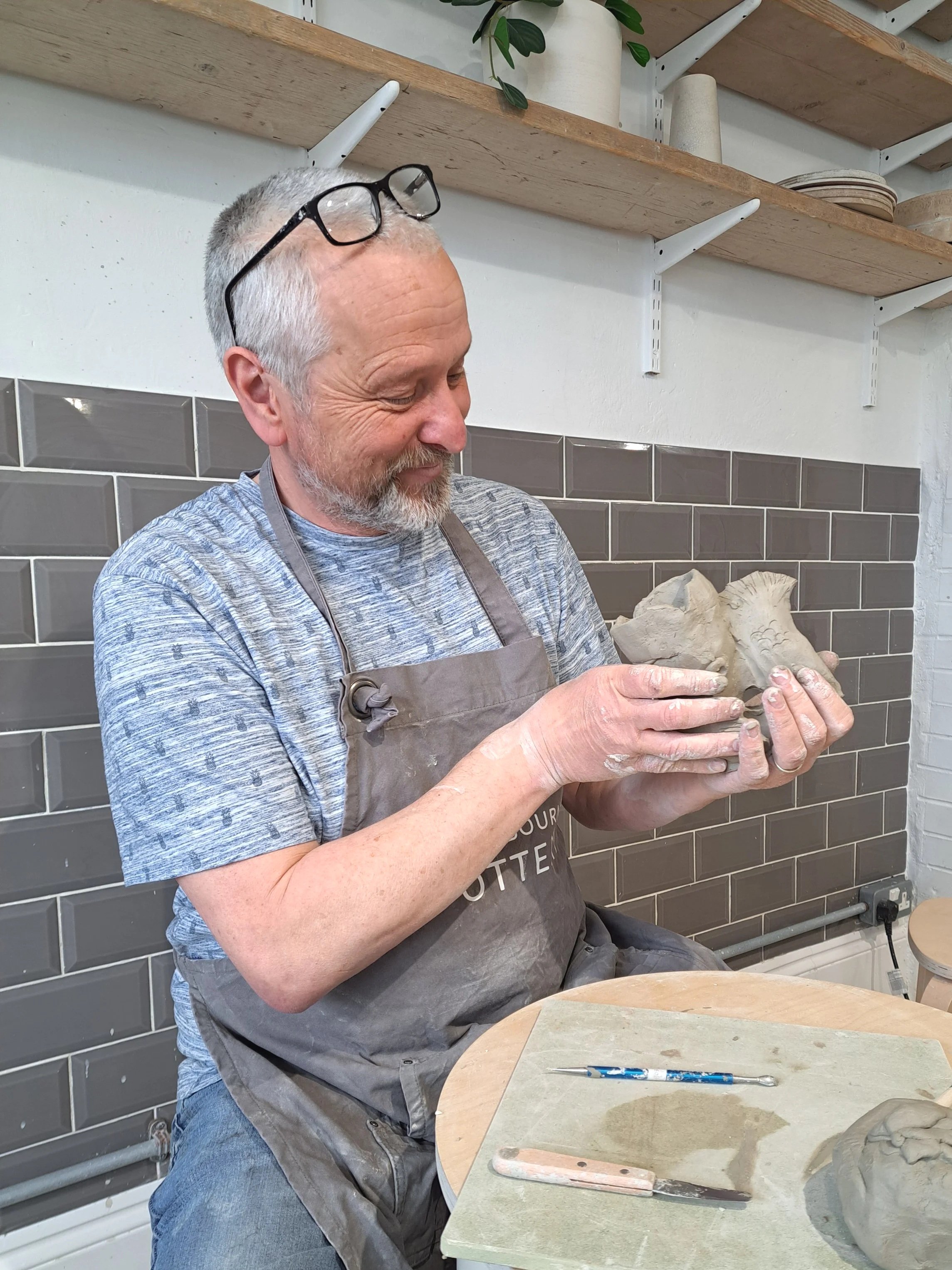 A man with glasses on his head and a beard, wearing a gray apron, is holding a piece of clay sculpture in a ceramics studio. There are tools on the table in front of him and shelves with pottery and plants in the background.