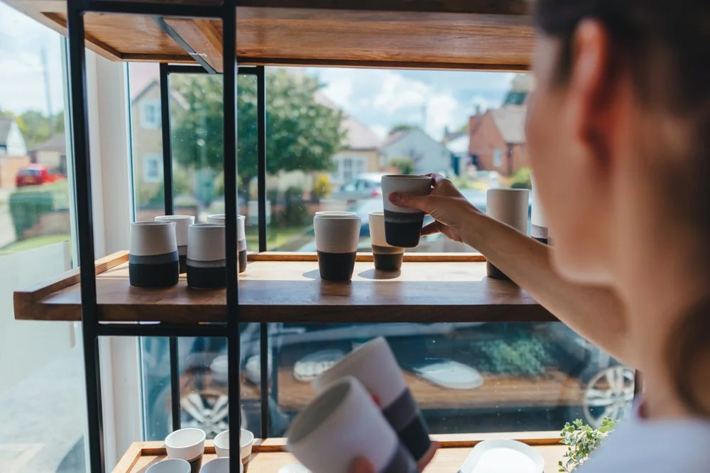 A woman selecting a ceramic cup from a wooden display shelf near a window with a suburban neighborhood view.
