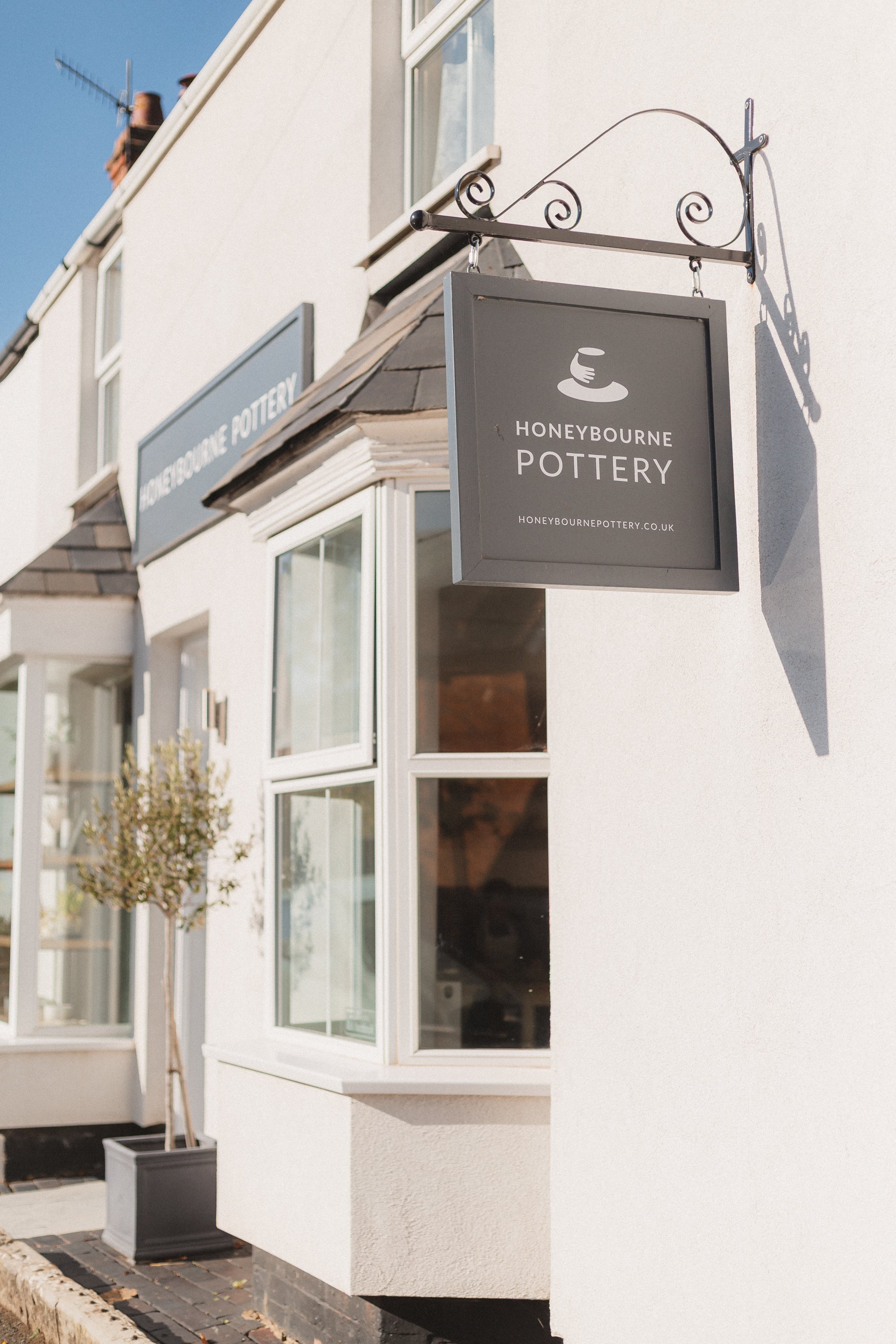Exterior view of a pottery shop named Honeybourne Pottery, with a hanging sign and bay windows, and a tree in a pot outside.