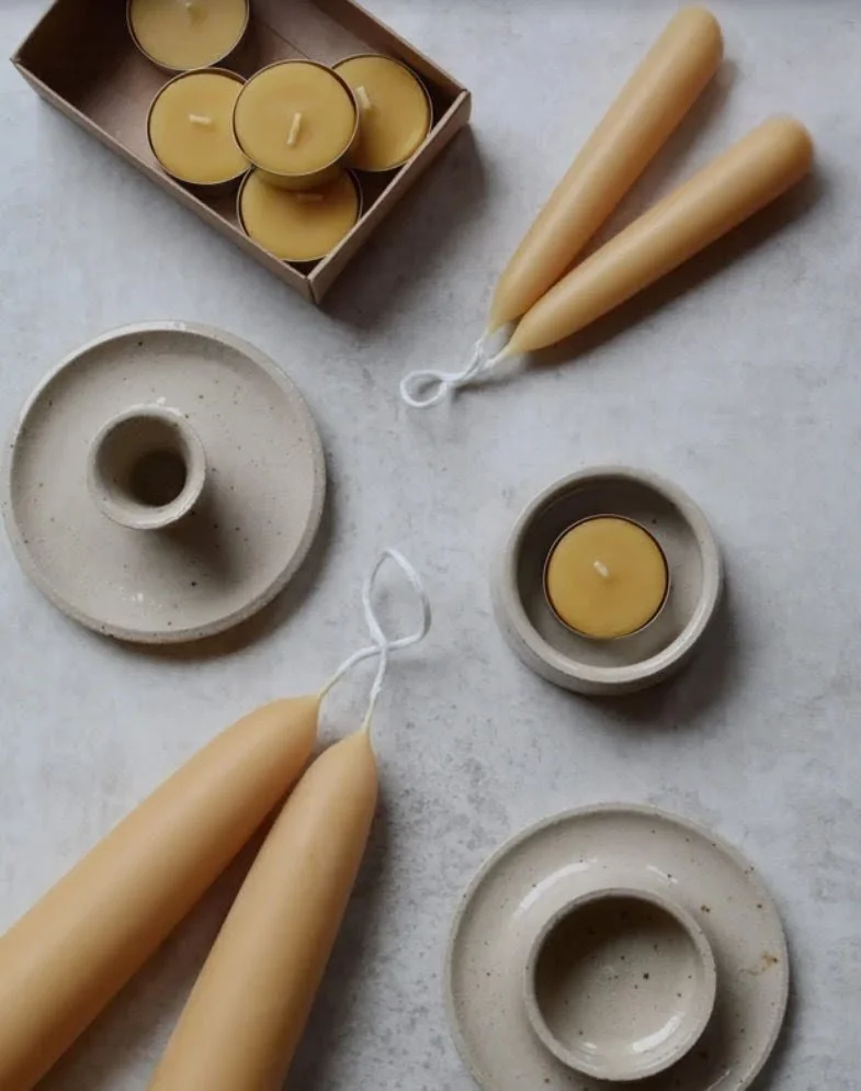 Cream-colored candles, some in small ceramic holders, with a box of tealight candles and two cream-colored taper candles on a light gray surface.